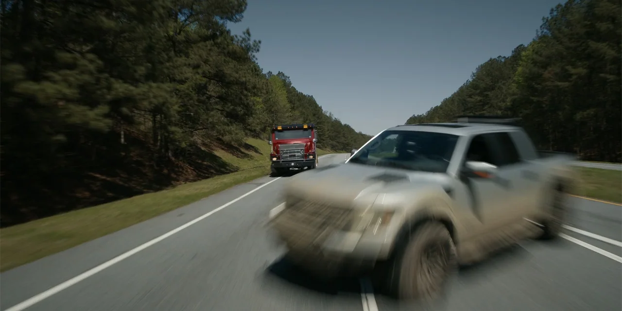 The Pickup (2025) movie still: wide — A two-lane highway passes through a rural landscape lined with pine trees under blue skies. A blurred silver…