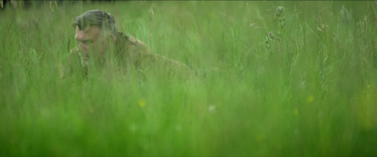 The Feast (2021) movie still: medium — A field of tall, thick green grass extends across the frame. An older man lies partially obscured within the…