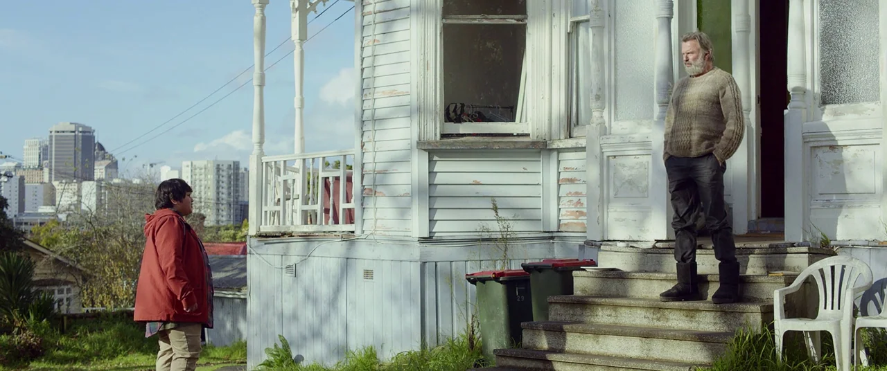 Hunt for the Wilderpeople (2016) movie still: wide — A wooden house with peeling paint features a porch overlooking a distant city skyline. An older…