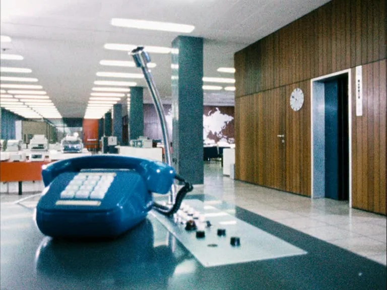 Making of: World On A Wire (1973) movie still: closeup — A spacious 1970s-style open office features wooden wall paneling and a world map mural. A…