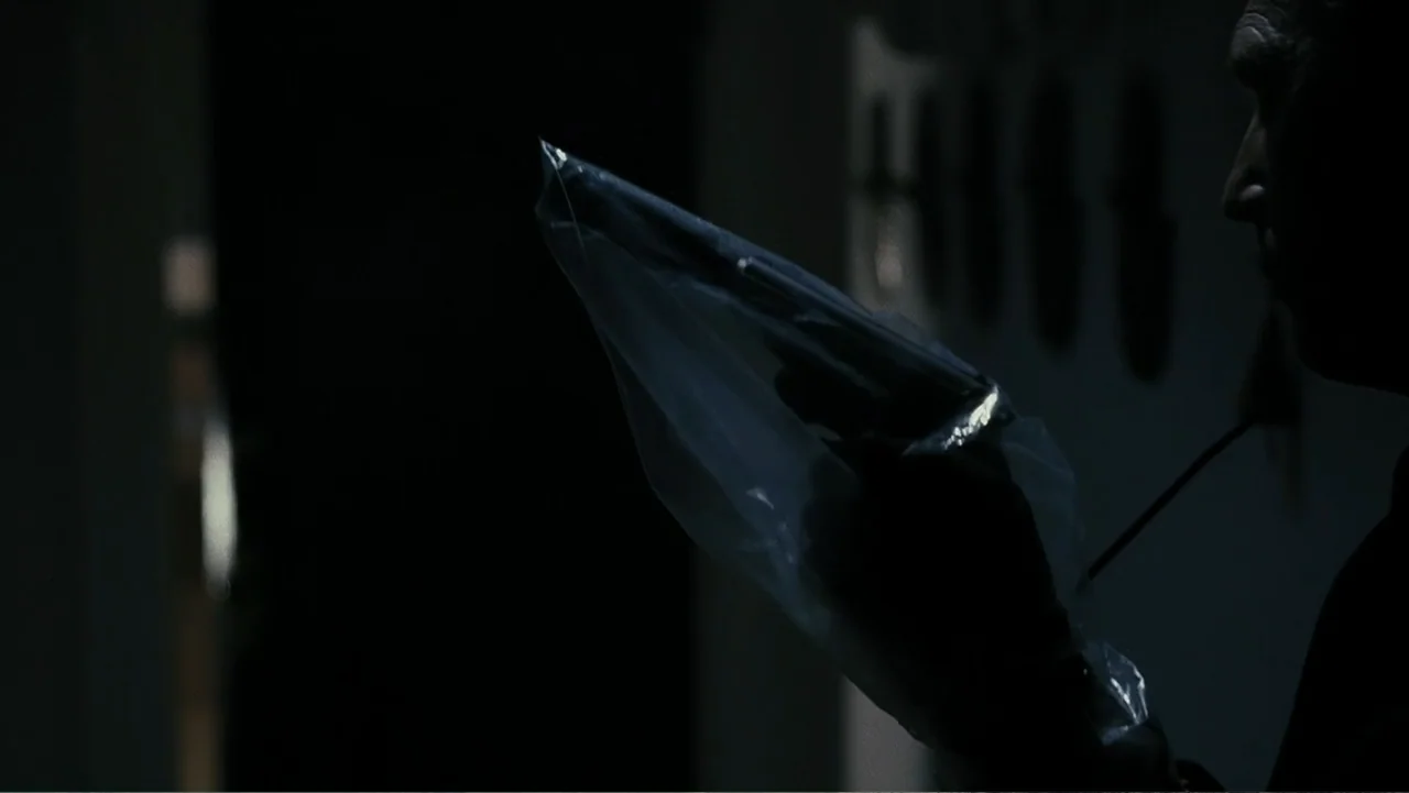 Mr. Brooks (2007) movie still: closeup — A dark, dimly lit room contains a person seen in silhouette from the side. A man holds a handgun tightly…