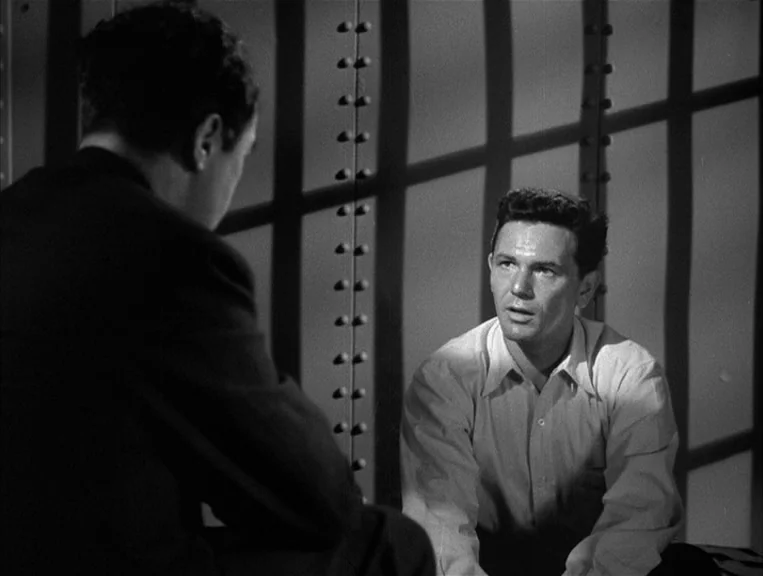 The Postman Always Rings Twice (1946) movie still: medium — A spartan room features a metal wall with vertical, riveted beams. One man in a light…