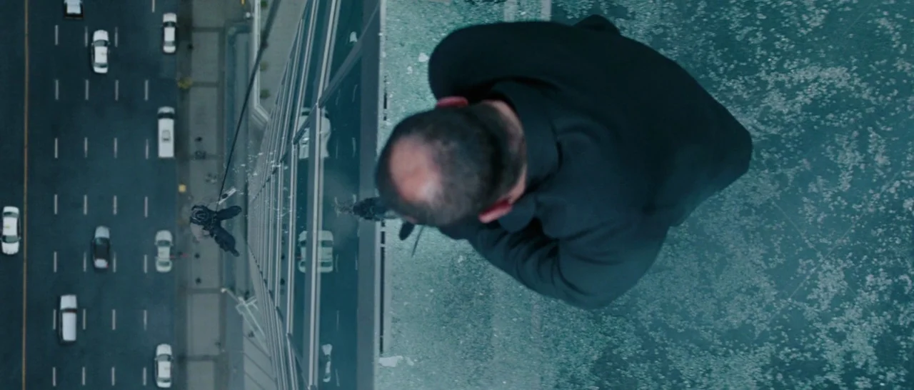 The A-Team (2010) movie still: wide — A man is positioned on the exterior wall of a glass skyscraper overlooking traffic. A second person is…