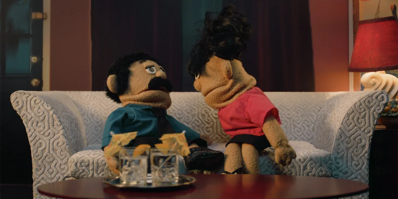 Música (2024) movie still: medium — A puppet man and a puppet woman sit together on a patterned fabric sofa. The figures are positioned facing each…