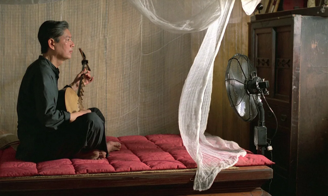 The Scent of Green Papaya (1993) movie still: medium — A man sits on a low wooden frame bed covered with a red mattress. White sheer mosquito…