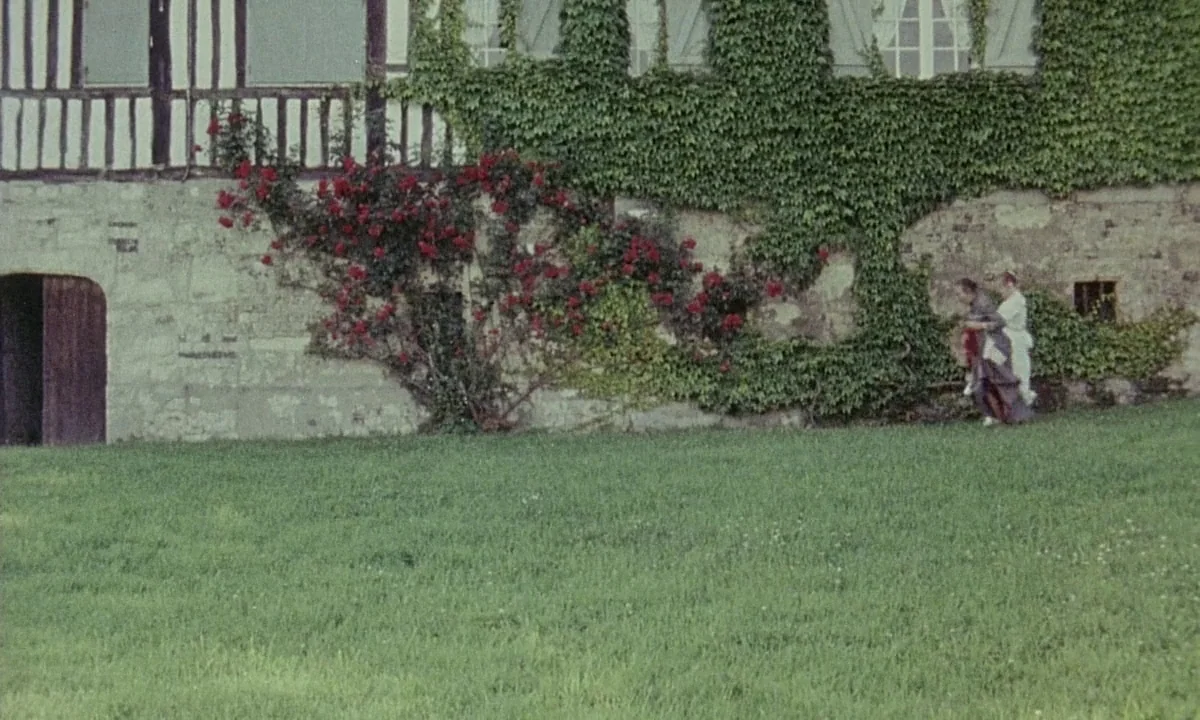 Immoral Tales (1974) movie still: wide — A stone building wall covered in thick ivy and climbing red roses rises above grass. Two women in vintage…