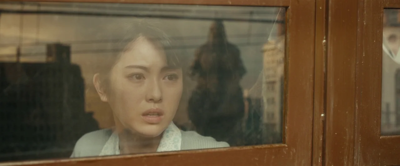 Godzilla Minus One (2023) movie still: closeup — A woman looks through a train window at a blurred cityscape with overhead lines. She presses…