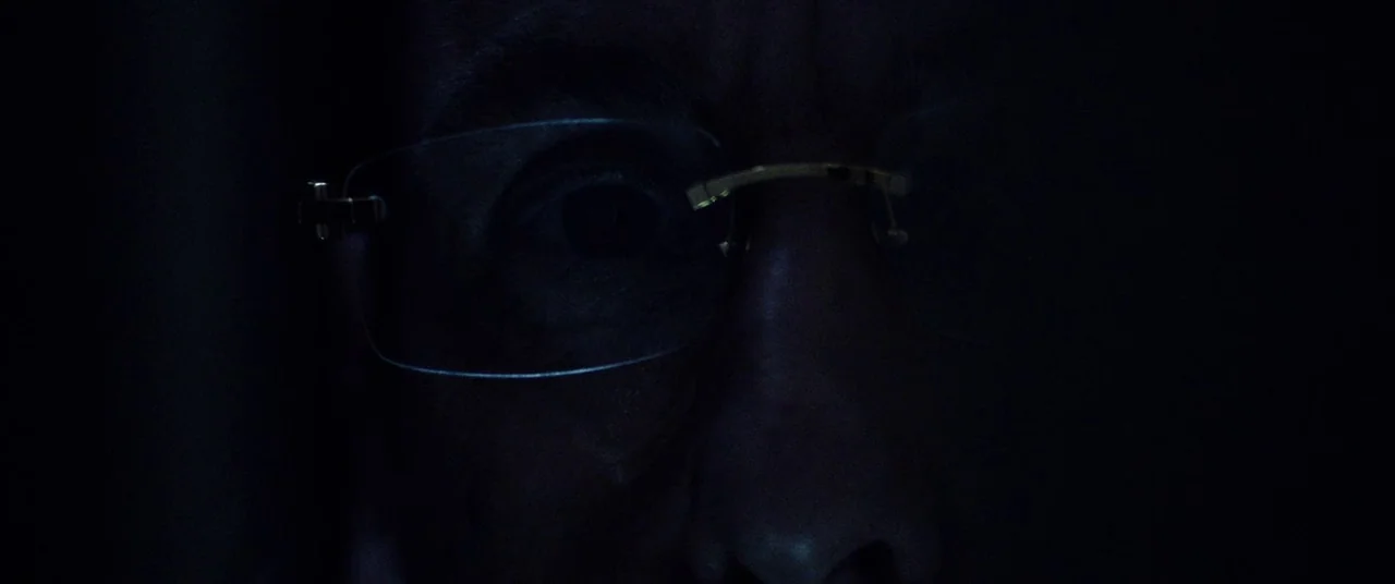 Uncut Gems (2019) movie still: closeup — A dark space obscures the environment and most of the subject's face. The subject wears wire-rimmed glasses…