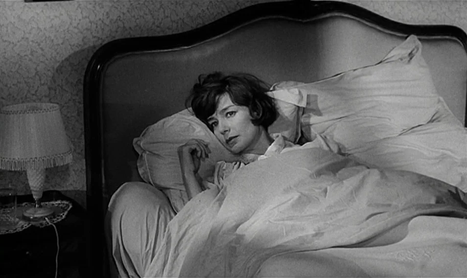 Thérèse Desqueyroux (1962) movie still: medium — A dark bedroom features a woman resting in a bed with a large upholstered headboard. The subject…