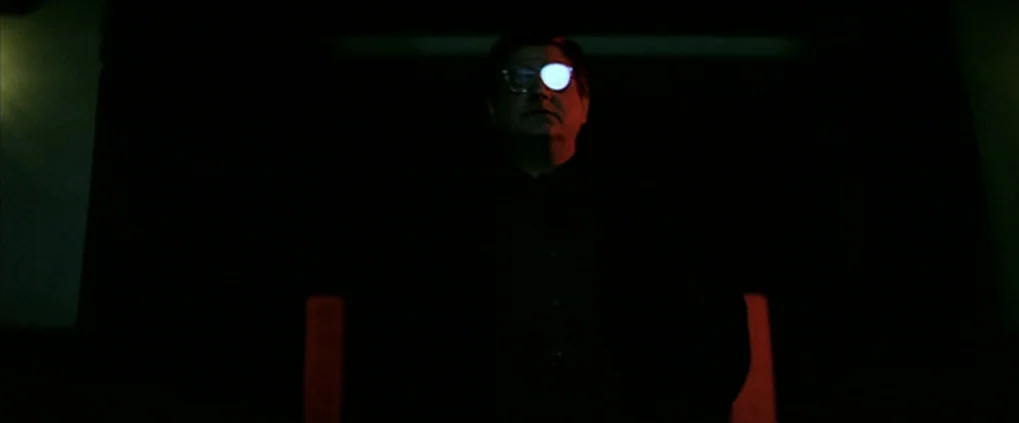 Anguish (1987) movie still: medium — A dark space shows a man lit primarily by a bright light on his face. He is wearing glasses and looking…