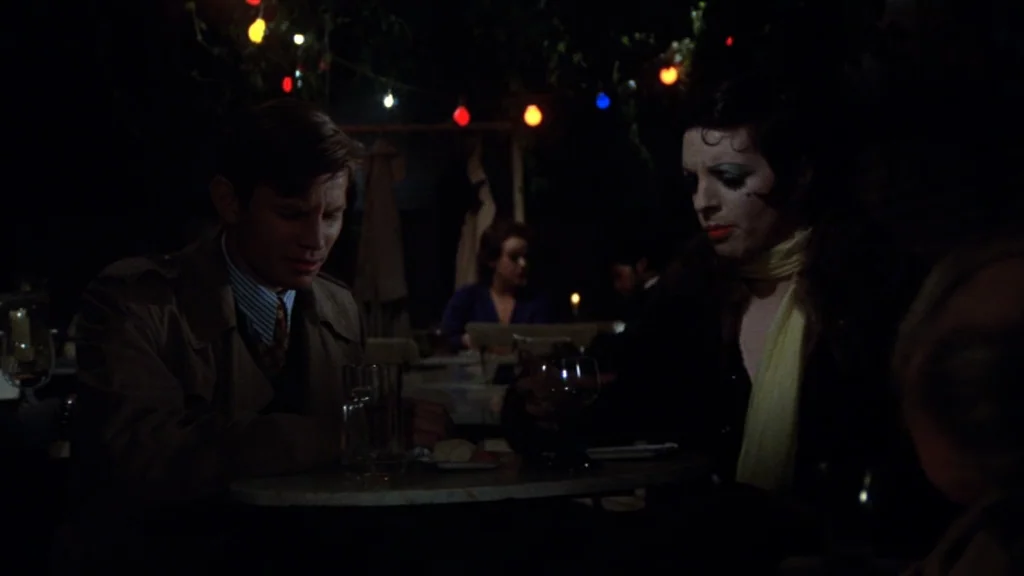Cabaret (1972) movie still: medium — An outdoor cafe area at night with colored string lights hanging above tables. A man and a woman sit closely…
