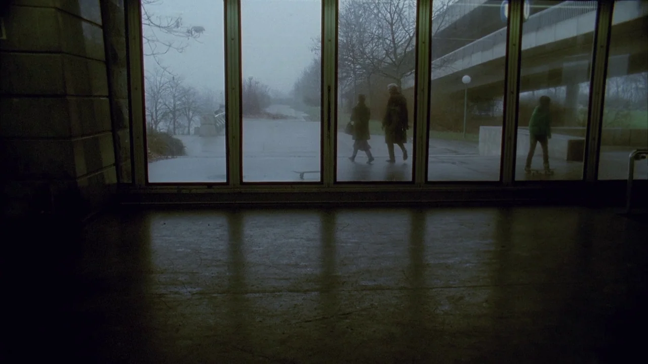 Museum Hours (2012) movie still: A somber, observational shot looking out from a dark, austere interior through a vast window wall. Outside, the…