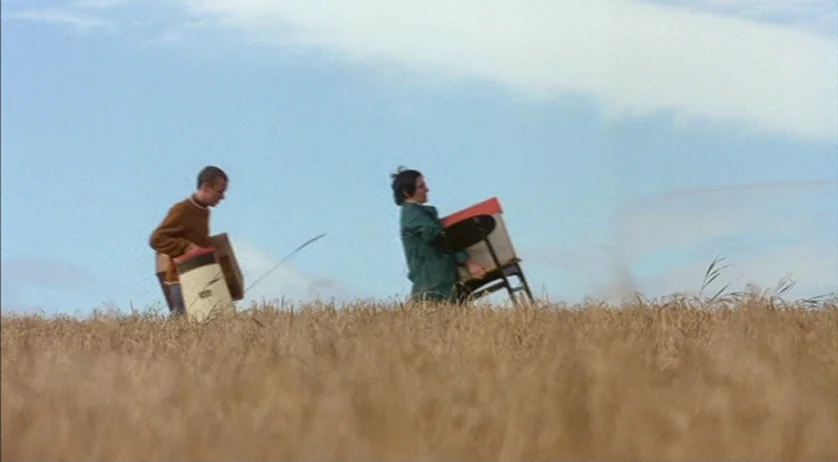 Ratcatcher (1999) movie still: Two figures traverse a vast, golden wheat field, carrying objects as they move across an expansive landscape. The…