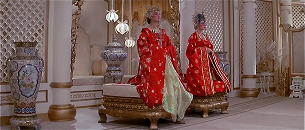 Big Trouble in Little China (1986) movie still: Two figures in ornate, vibrant red silk robes sit motionless on gilded platforms within a palatial…