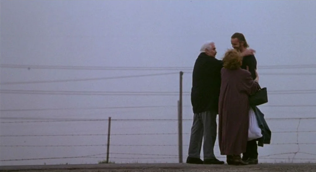 The Crossing Guard (1995) movie still: fullbody — A flat, barren landscape under a heavy grey sky with a simple barbed wire fence. Three people…