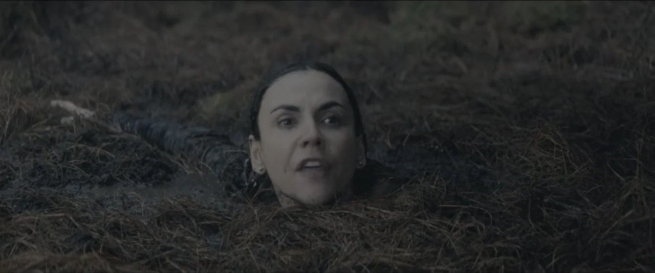 Quicksand (2023) movie still: closeup — A dark muddy patch covered in brown pine needles contains a single person. A woman with dark hair emerging…