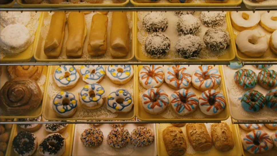 Youth In Revolt (2009) movie still: medium — A multi-tiered glass display case holds various rows of colorful glazed donuts and pastries. White…