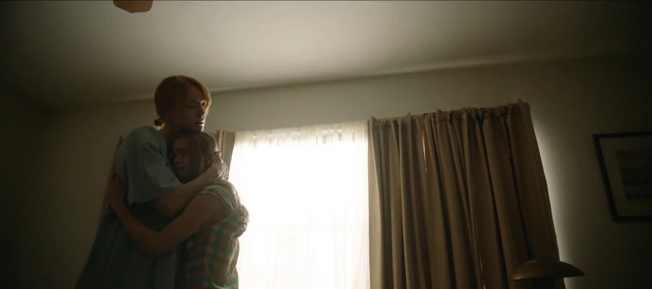 Fuzzy Head (2023) movie still: medium — A spare room features a window covered by beige curtains with a single framed picture. An adult woman stands…