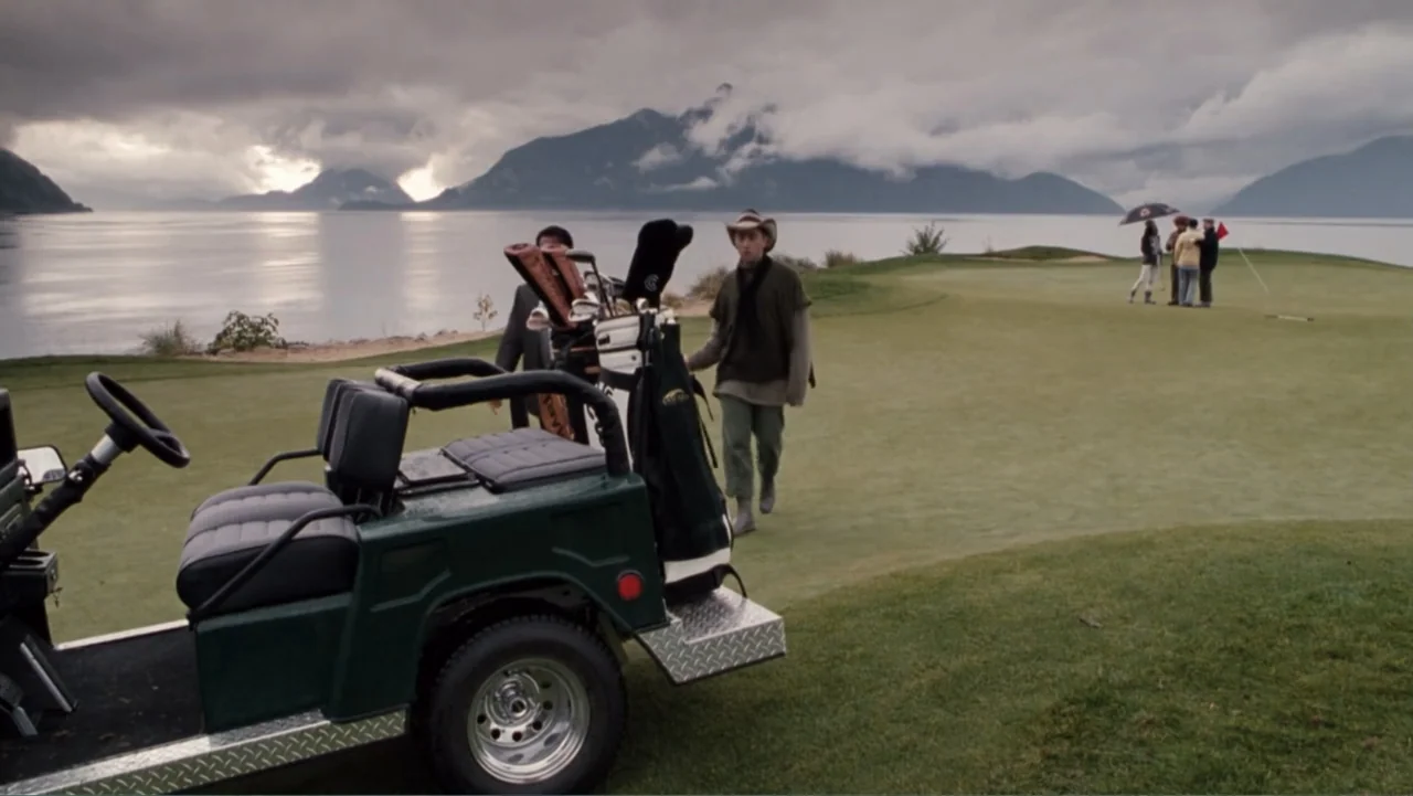 Get Carter (2000) movie still: wide — A coastal golf course features rolling green turf bordered by a large mountain lake. A lone man walks across…