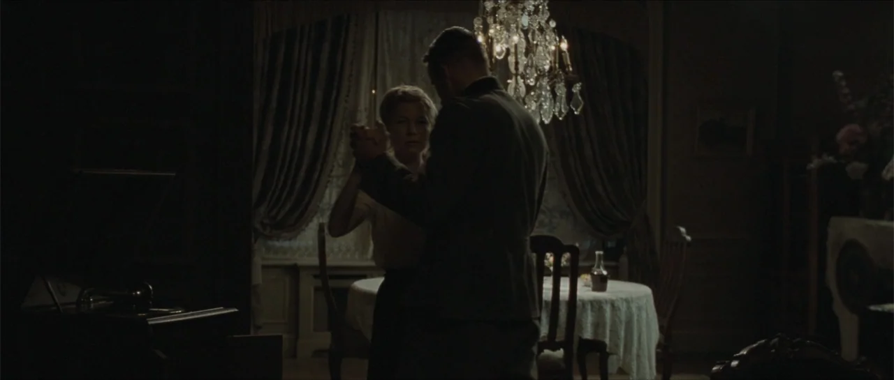 Suite Française (2015) movie still: medium — A dimly lit room contains a draped round table and a hanging crystal chandelier. A man and a woman…
