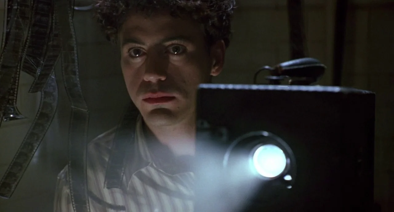 Chaplin (1992) movie still: closeup — A dark space with photographic film strips hanging in the foreground. A young man watches a light emanating…