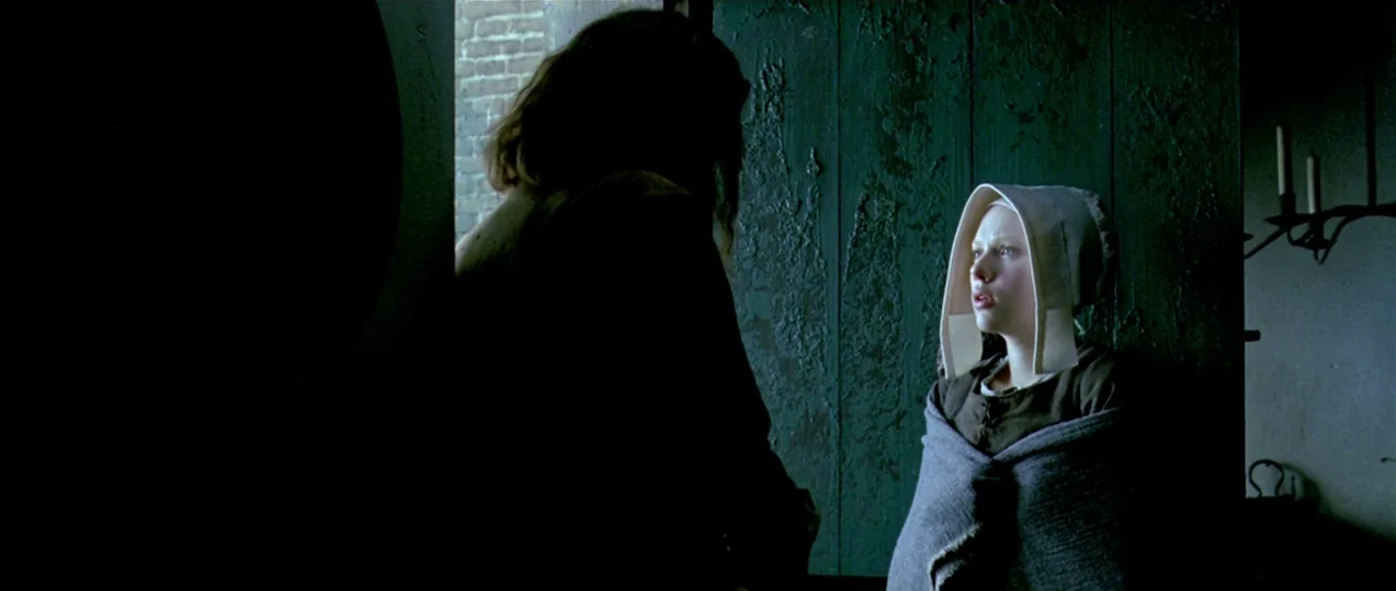 Girl with a Pearl Earring (2003) movie still: medium — A dark cottage room features a green wooden door and a single window. A girl in a white cap…