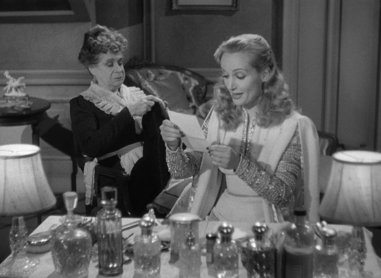 To Be or Not To Be (1942) movie still: medium — An interior space features a vanity covered in glass perfume bottles and lamps. A young woman reads…