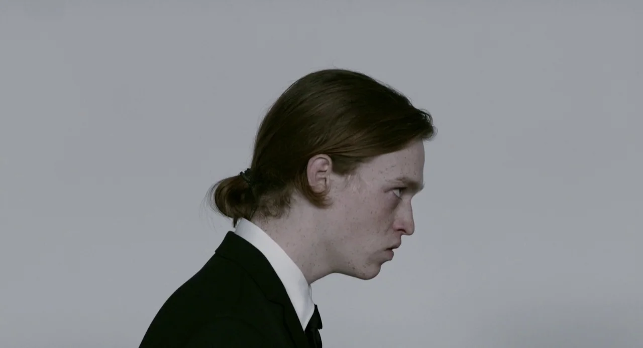 Antiviral (2012) movie still: closeup — A man in a formal black suit stands against a plain light gray background. The subject is positioned in…
