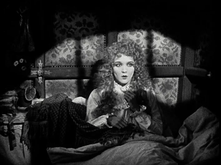 The Man Who Laughs (1928) movie still: medium — A woman sits upright in a bed against a floral patterned wall in shadow. She clutches her hands to…
