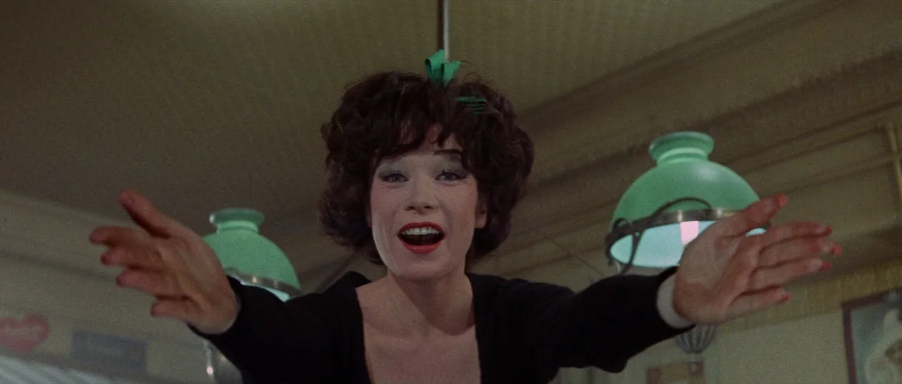 Irma La Douce (1963) movie still: medium — A woman stands in a venue featuring distinctive green-shaded glass pendant lamps. She extends her arms…