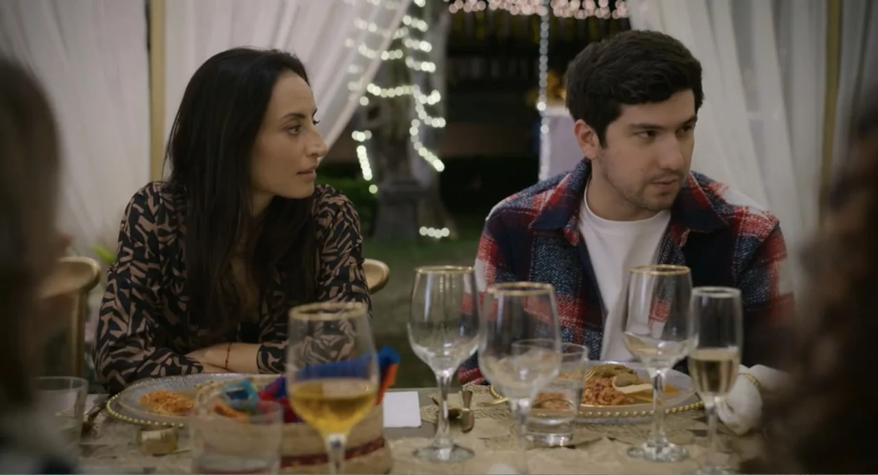 La posada (2023) movie still: medium — An outdoor dining area is decorated with white curtains and string lights at night. A young man and woman sit…