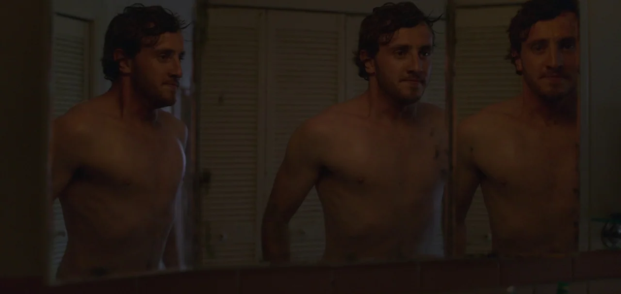 Foe (2023) movie still: medium — A dimly lit residential bathroom features white louvered closet doors in the background. A shirtless man stands in…