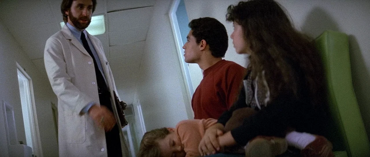 The Entity (1982) movie still: medium — A hospital corridor with off-white walls and green plastic chairs lining the wall. A doctor standing in a…