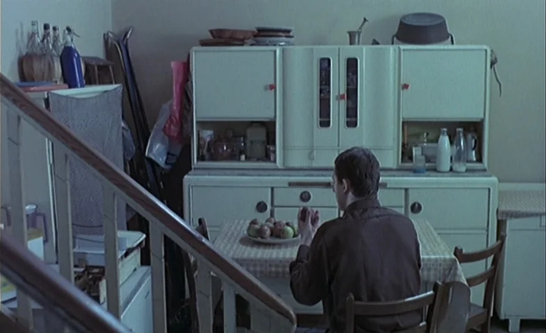 Blind Chance (1987) movie still: medium — A vintage dining area features a large white cabinet and a small wooden table. A man in a brown shirt sits…