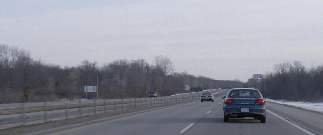 The End of the Tour (2015) movie still: wide — A long stretch of multi-lane highway cuts through a dense forest of winter trees. Two cars travel…