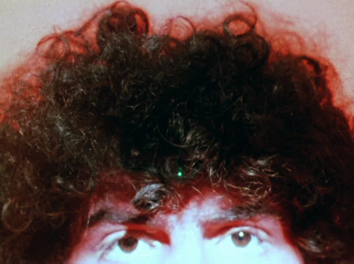 Lucifer Rising (1972) movie still: closeup — The frame shows a close view of a person with dark curly hair. A red light illuminates the edges of the…