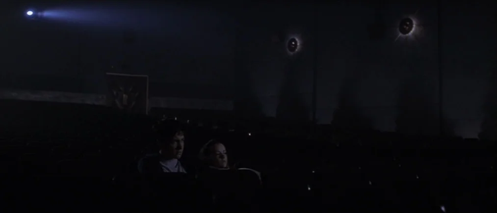Donnie Darko movie still: wide — A dimly lit, empty movie theater auditorium with rows of dark, tiered seats. A young couple sits together in the…