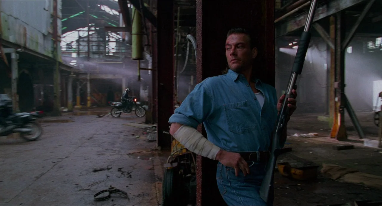Hard Target (1993) movie still: medium — An abandoned industrial facility with rusted support beams and dust-filled air. A man holding a shotgun…