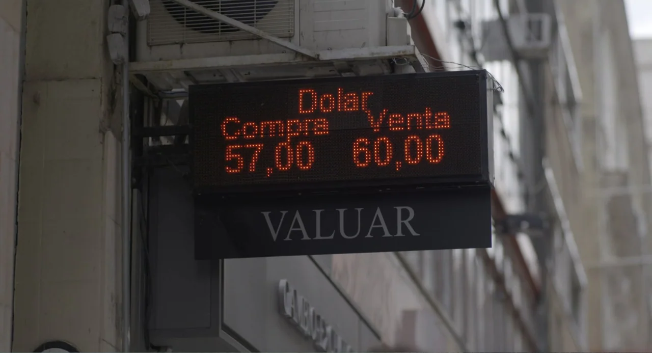 An Odd Turn (2024) movie still: closeup — A dark rectangular LED sign displaying currency exchange rates hangs on a gray building wall. The sign…