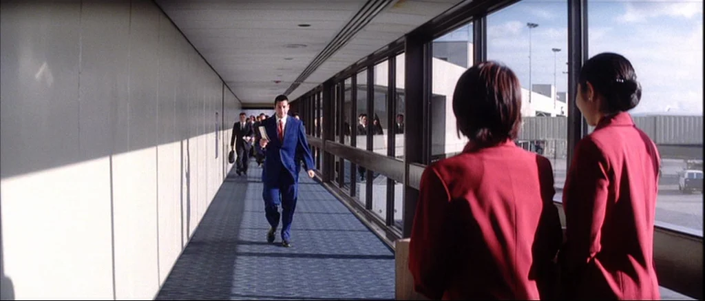 Punch Drunk Love (2002) movie still: fullbody — An airport jet bridge features large glass windows overlooking a tarmac area in sunlight. A man in a…