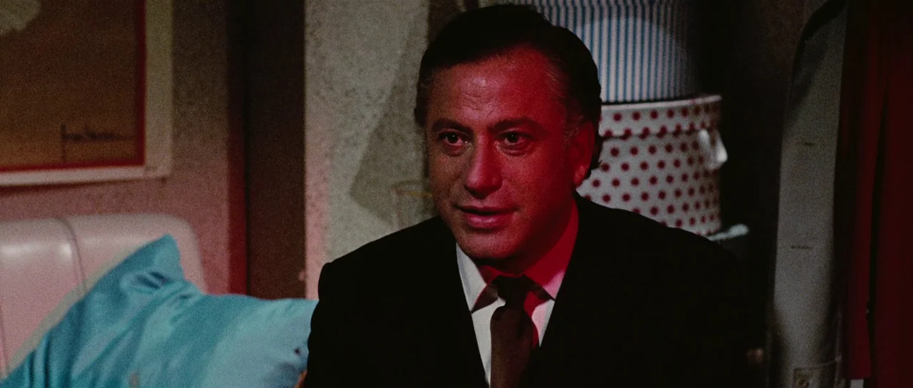 Death Walks On High Heels (1971) movie still: closeup — A dimly lit room contains patterned storage boxes stacked in the background. A man in a dark…