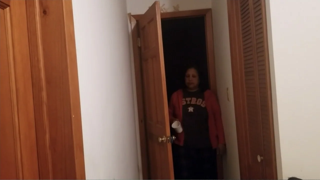 Covid Diaries NYC (2021) movie still: medium — A woman stands in a wooden doorway connecting two interior rooms. She holds a white mug while facing…