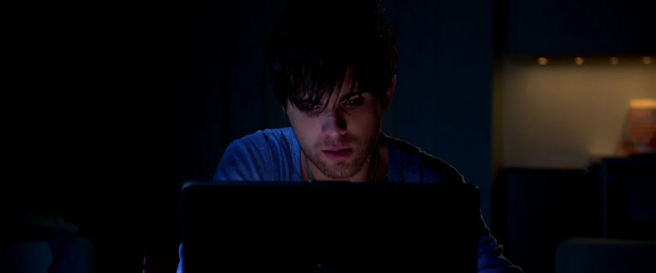 Kaboom (2010) movie still: medium — A dark, dimly lit indoor space with blurred background details suggesting a home setting. A young man sits in…