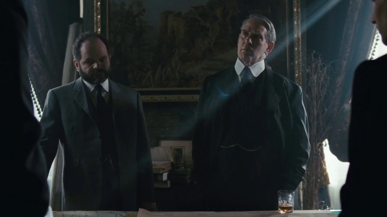 Childhood of a Leader (2015) movie still: medium — A dimly lit room with wood-paneled walls and a large framed painting in background. Two men in…