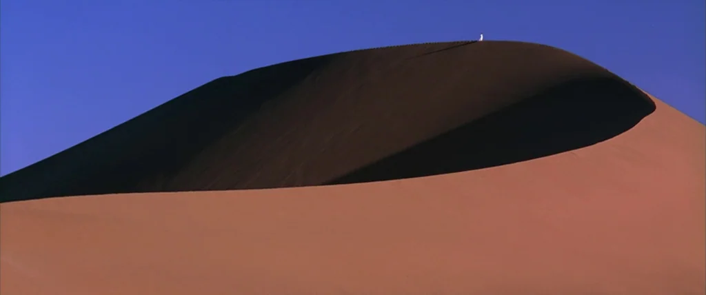 The Cell (2000) movie still: establishing — A vast, smooth sand dune stands under a clear, bright blue sky. The curve of the dune creates a…