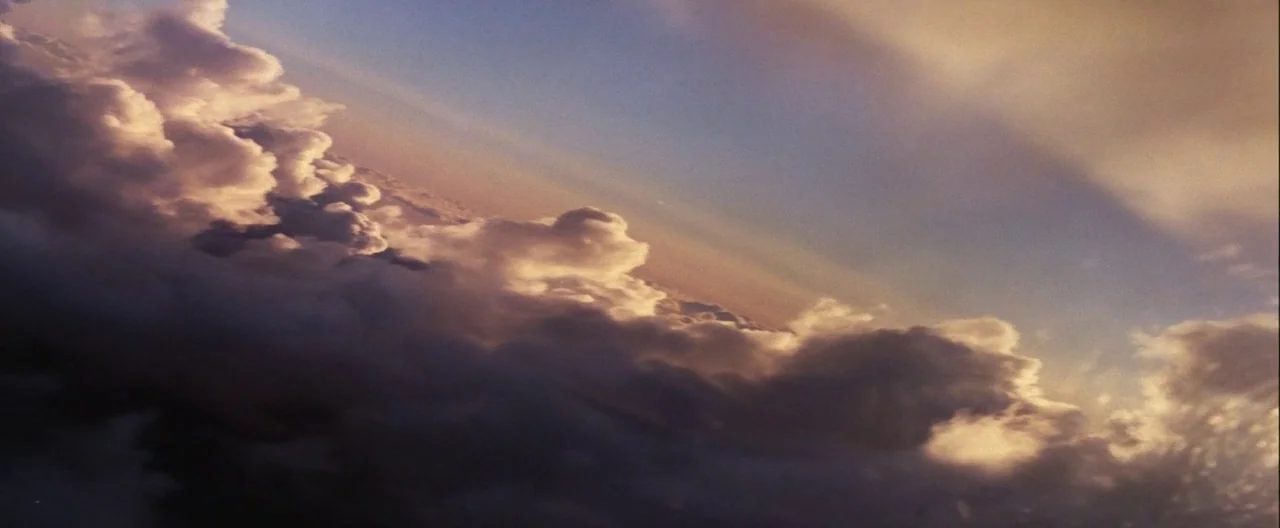 Grease (1978) movie still: establishing — Large cumulus cloud formations fill the frame at high altitude. Sunlit edges contrast against deeper grey…