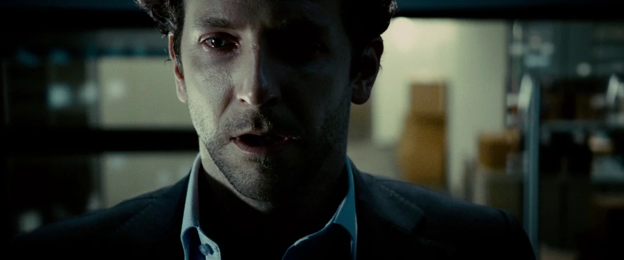 Limitless (2011) movie still: closeup — A man stands in a dimly lit, narrow passage with blurred items in the background. He is centered in the…