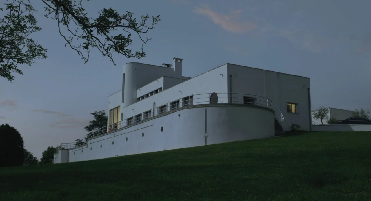 Holy Motors (2012) movie still: establishing — A stark white modernist villa with curved walls sits on a grassy hill at dusk. A branch hangs in the…