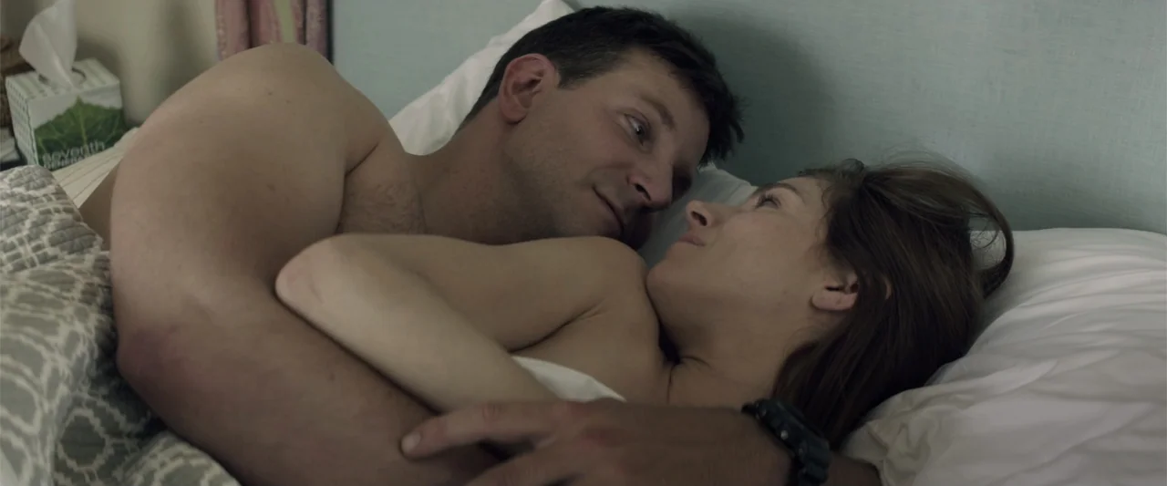 American Sniper (2014) movie still: medium — A modern bedroom with a light blue padded headboard and patterned grey bedding. A man and a woman are…