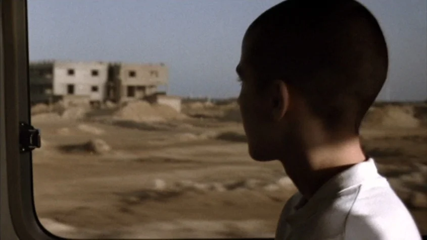 Benny’s Video (1992) movie still: medium — A view through a train window shows an arid landscape with a half-built concrete structure. A young man…